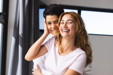 Portrait of happy biracial lesbian couple embracing in sun by window at home