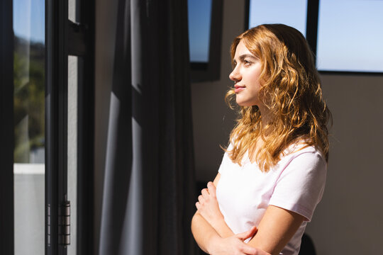 Thoughtful biracial woman looking through window at home, smiling on sunny day, copy space