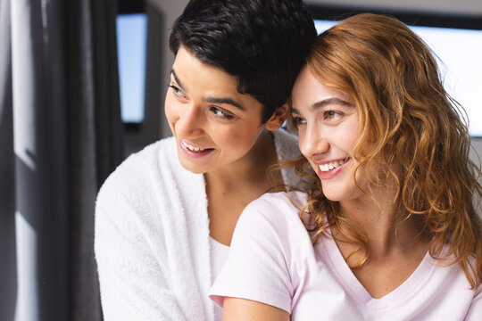 Happy biracial lesbian couple embracing in sun looking out of window at home