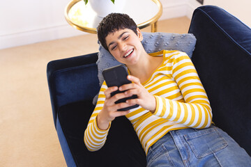 Happy biracial woman lying on sofa using smartphone at home and smiling - Powered by Adobe