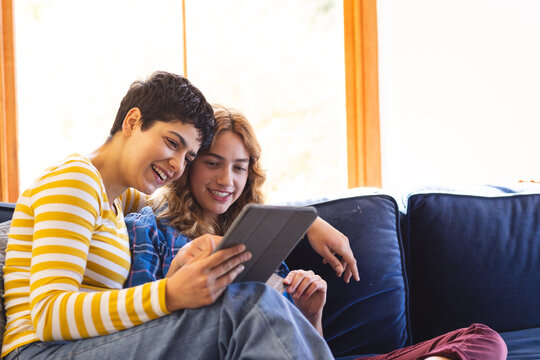 Happy biracial lesbian couple relaxing on couch using tablet together, copy space