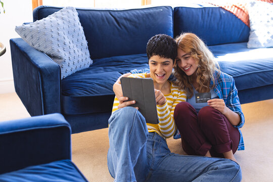 Happy biracial lesbian couple sitting on floor shopping using tablet and credit card, copy space