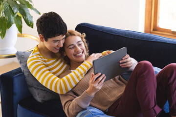 Happy biracial lesbian couple embracing on couch using tablet at home