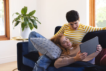 Happy biracial lesbian couple relaxing on couch together using tablet at home, copy space