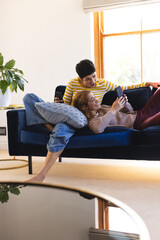 Happy, surprised biracial lesbian couple relaxing on couch together using smartphones, copy space