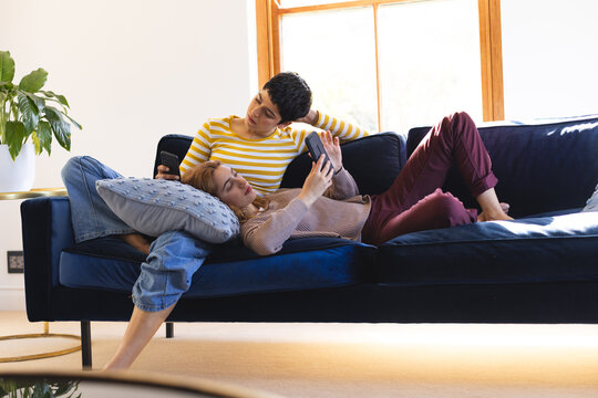 Happy biracial lesbian couple relaxing on couch together using smartphones at home