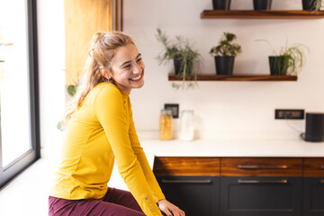 Happy biracial woman sitting on countertop smiling in kitchen, copy space
