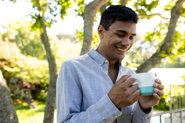 Happy biracial man drinking coffee and standing on balcony at home