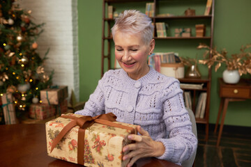 Obraz na płótnie Canvas Portrait of happy senior female in cardigan looking and touching gift box with ribbon, ready to unpack and trying to guess about its content on background with lighted and decorated christmas tree
