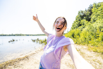 Fototapeta na wymiar In the embrace of the great outdoors, a happy young woman snaps a selfie by the lakeside, her vibrant smile capturing the essence of a sun-kissed day. This snapshot is a celebration of health