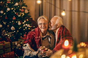 A festive senior couple is sitting on the floor at christmas and new year's eve, kissing, hugging...
