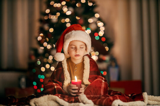 A cute boy is praying with candle in his hands while sitting at home on christmas eve.