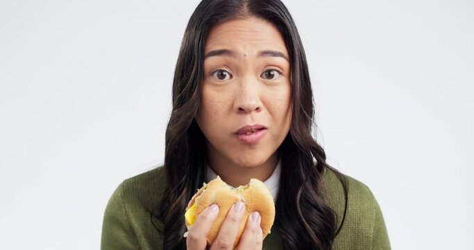 Young woman, eating burger and studio with face, french fries or comic for greed by white background. Japanese girl, student and fast food with bad choice for health, sandwich or portrait with chips
