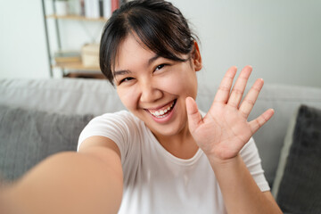 Young Asian woman using smartphone for online video conference call with friends waving hand making...