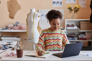 Young African American craftswoman watching online educational video and making notes in copybook while sitting by workplace in craft shop