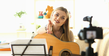 Fototapeta na wymiar Playing Music Lessons Recording for Social Network. Beautiful Female Blogger Performer Holding Guitar Extending Hand to Camcorder. Smiling Musician Beauty Broadcast Online Musical Workshop