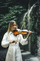 Charming Caucasian woman playing violin with closed eyes near waterfall. Music and art concept. Female with long blond hair wearing white dress. Water splash. Banyu Wana Amertha waterfall