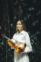 Portrait of beautiful Caucasian woman holding violin in tropical forest. Music and art concept. Female wearing white dress. Background of lush green tropical leaves. Copy space. Vertical layout.