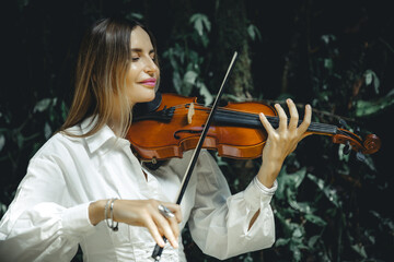 Close up portrait of smiling Caucasian woman playing violin in tropical forest. Closed eyes. Music and art concept. Female wearing white dress in nature. Background of lush green tropical leaves.