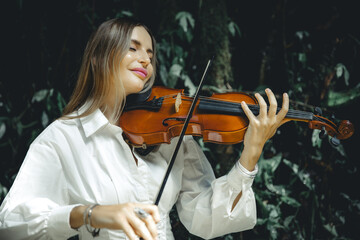 Close up portrait of smiling Caucasian woman playing violin in tropical forest. Closed eyes. Music...
