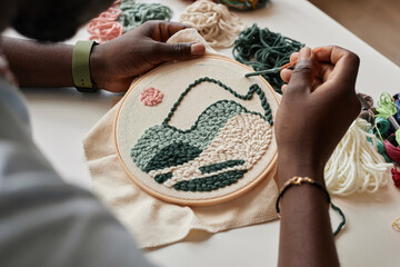 Close-up of young creative male artisan creating embroidery on piece of white canvas with special...