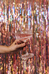 Persons hand holding glass of champagne wine over sparkling tinsel curtain. Foil fringe shimmer...