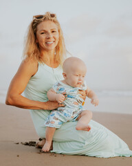 Young Caucasian mother holding infant baby boy, spending time on the beach. Sitting on the sand. Happy smiling mum with little son. Summer vacation. Family concept. Sunset time. Seminyak, Bali