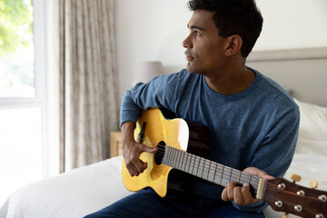 Biracial man sitting on bed playing guitar at home