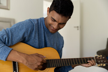 Happy biracial man sitting on bed playing guitar at home - Powered by Adobe