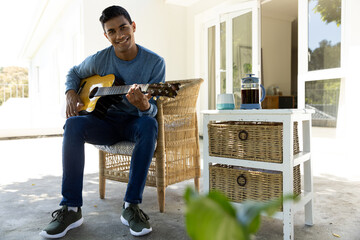 Portrait of happy biracial man sitting on balcony playing guitar at home, copy space - Powered by Adobe