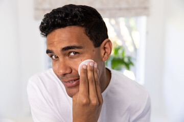 Fototapeta na wymiar Happy biracial man cleansing face with cotton pad in bathroom at home