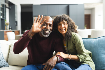 Happy mature diverse couple having video call, waving on couch in sunny living room