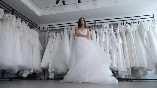 Slow motion of a beautiful slender bride spinning in a wedding salon against the background of many white dresses hanging on a hanger. A happy woman tries on a wedding dress and is spinning. 