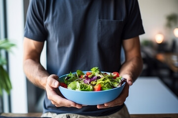 Man eat healthy lunch in modern interior, Unrecognizable profile male torso in blue t-shirt, hand with fork, near window with vegetable salad in bowl, diet food concept