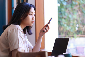pensive asian woman sitting near window with laptop on wood table holding smartphone looking away...