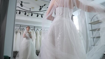 A happy young woman is trying on a wedding dress in a wedding salon, she is standing in front of a mirror and spinning. A long transparent veil flutters in different directions. A chic wedding image. - Powered by Adobe