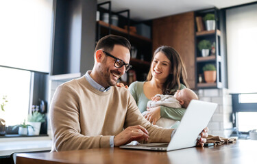 Young father works on laptop while his wife looks after child.Work from home parenting telecommuting - obrazy, fototapety, plakaty
