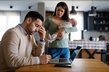 Stressful man trying to work on laptop with wife and children on background at home office.