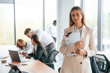 Young woman is holding documents in hands, standing. Group of office workers are together indoors
