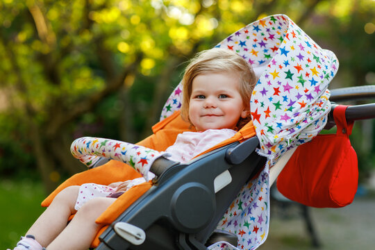 Portrait of little cute smiling toddler girl sitting in stroller or pram and going for a walk. Happy cute baby child having fun outdoors. Healthy daughter.