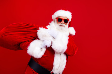 Portrait of satisfied optimistic santa claus in stylish sunglass holding bag with presents on shoulder isolated on red color background