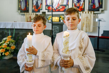Two little kids boys receiving his first holy communion. Happy children holding Christening candle. Tradition in catholic curch. Kids in a church near altar. Siblings, brothers in white gowns.
