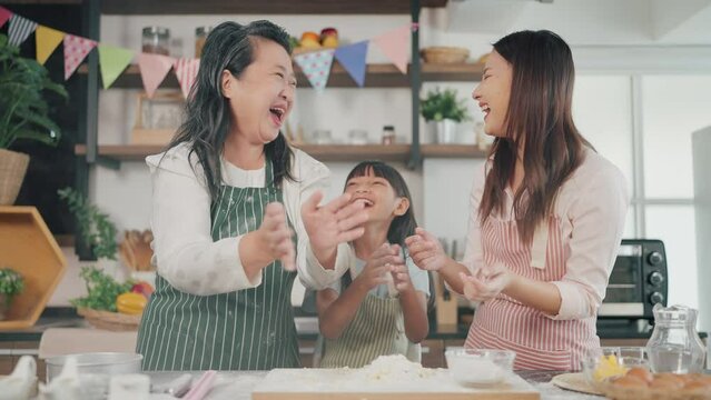 Cheerful Three Generations Asian Women In Apron Having Fun In Kitchen. Grandmother, Mother And Little Daughter Clapping Floury Hands Messy, Shouting And Laughing. Family And Cooking Concept