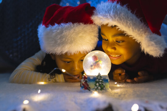 African american boy and girl lying and looking at illuminated christmas snow globe at home