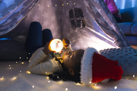 African american girl lying, wearing hat and playing with illuminated christmas snow globe at home - Powered by Adobe