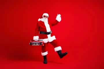  Full length photo of optimistic santa claus in sunglass waving palm say hello hold boombox dancing isolated on red color background © deagreez