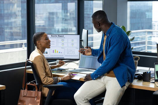 Happy african american male and female colleague looking at computer and talking at desk in office