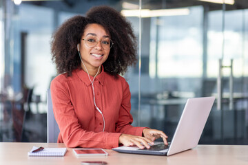 Portrait of young beautiful successful woman at workplace inside office, businesswoman in headphones smiling looking at camera, satisfied with achievement results programmer with laptop.