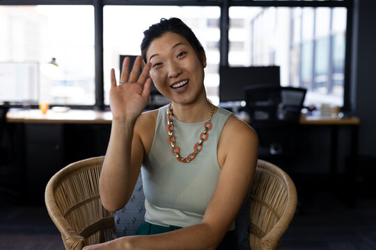 Happy asian casual businesswoman having video call waving from armchair in office