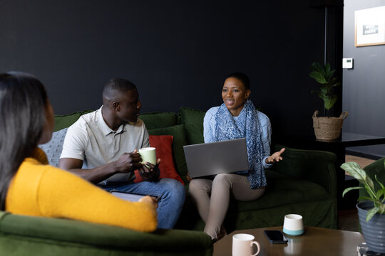 Happy diverse male and female colleagues with laptop having coffee and talking in office lounge